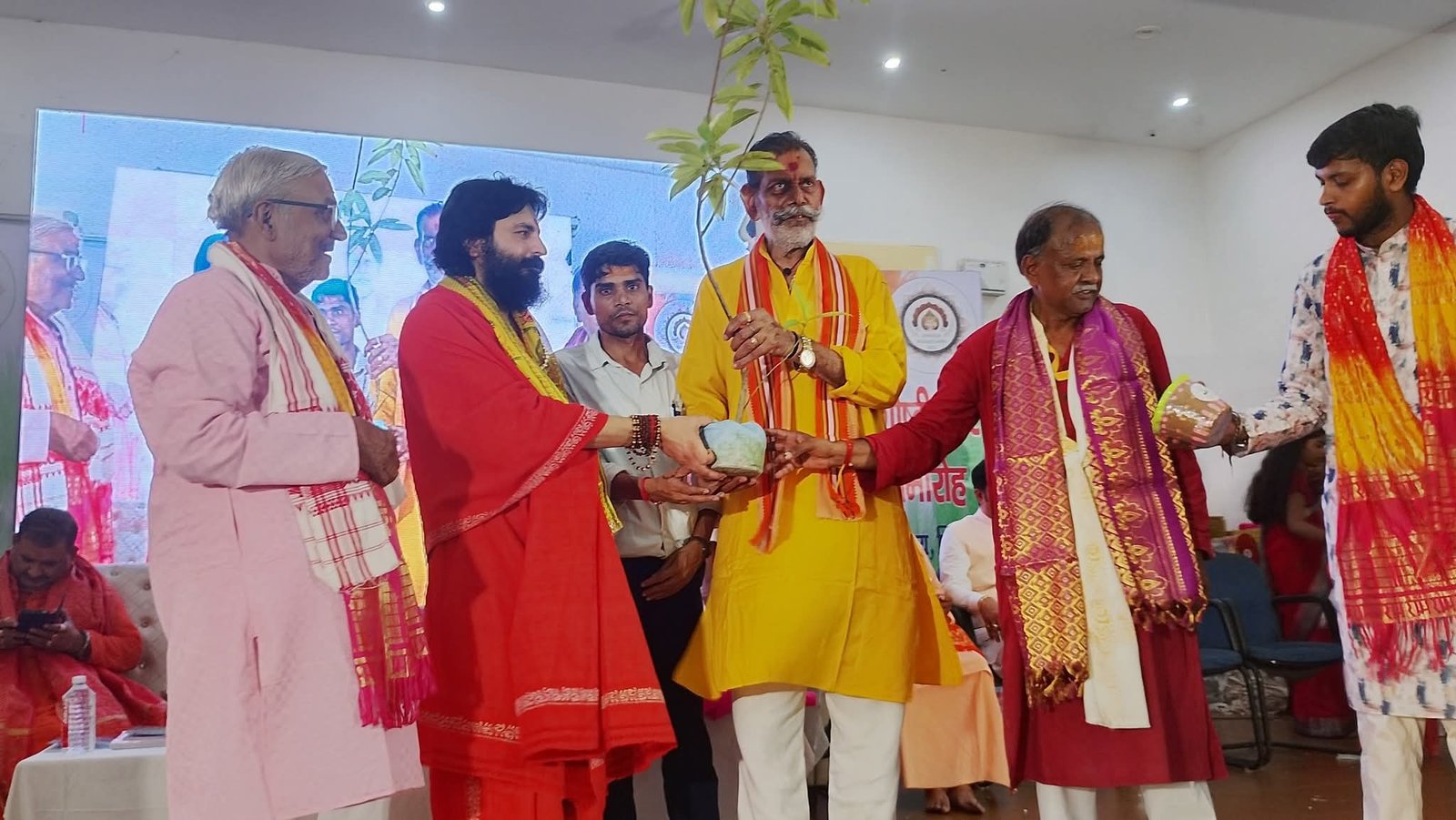 A man in a red robe presenting a potted sapling to a man in a yellow kurta on stage, with others in ceremonial scarves looking on