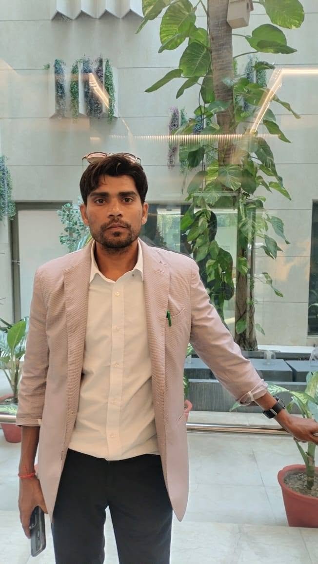 A man in a light blazer standing in a modern building atrium with plants and glass walls behind him