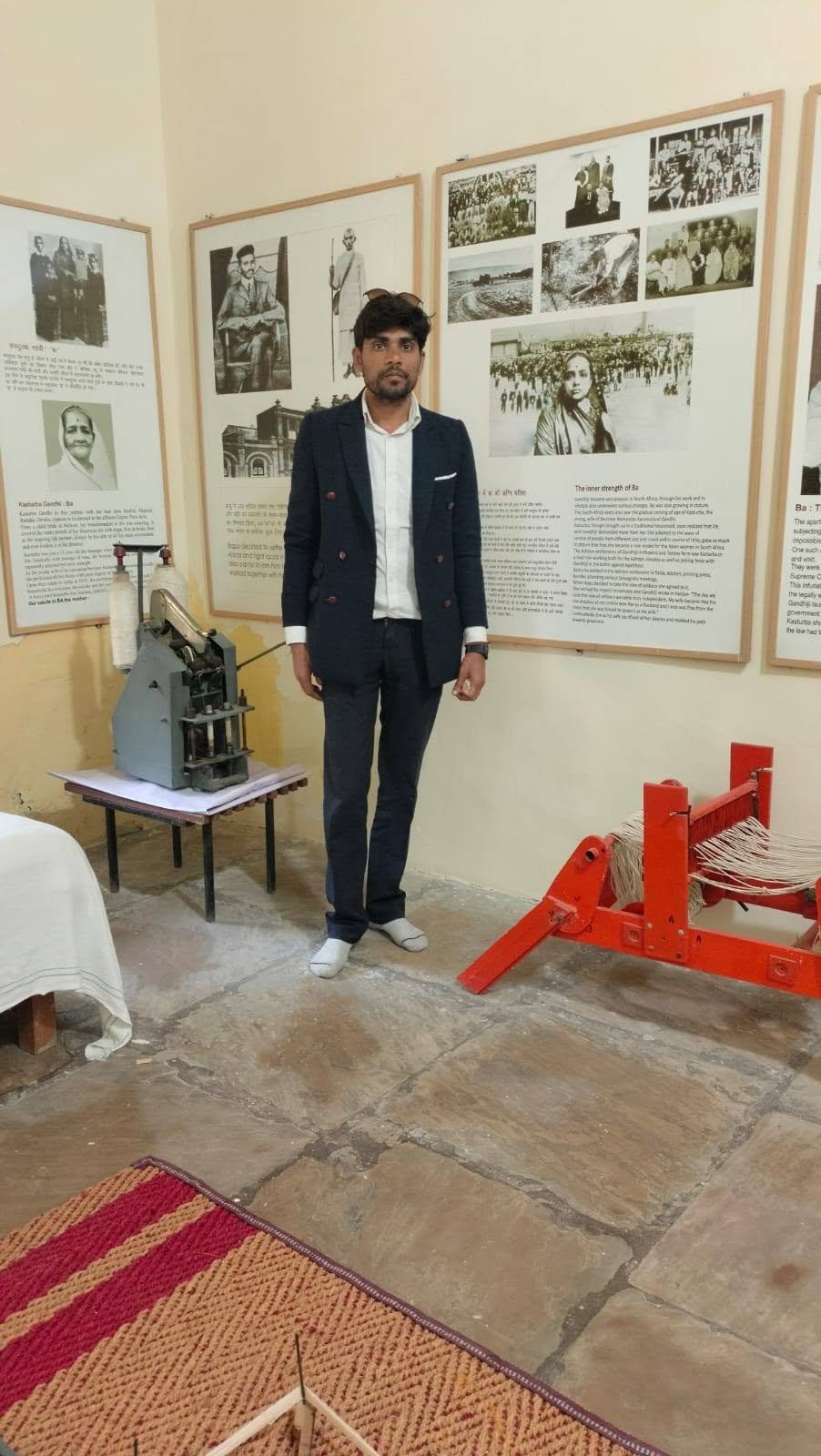 A man in a dark blazer standing in a museum room among historical photo displays and a spinning wheel exhibit