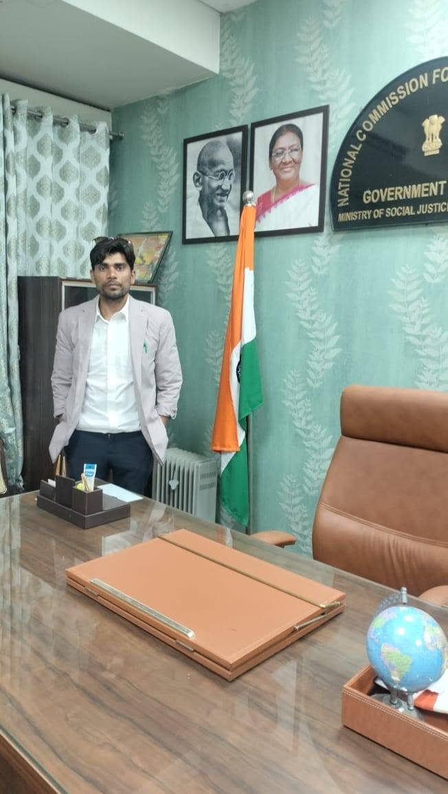 A man in a light blazer standing behind a desk in a government office beside an Indian flag on a stand